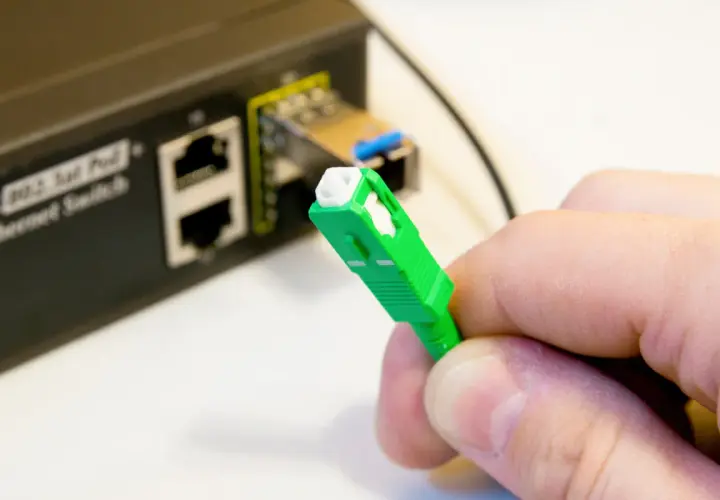 Dłoń trzyma zieloną wtyczkę światłowodową. Obok widać przełącznik Ethernet. Tak sprawdzisz, czy jest światłowód.