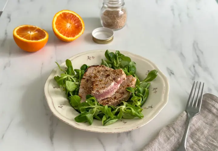 Tonno in padella con crosta di sesamo su letto di insalata fresca, accompagnato da spicchi d'arancia.
