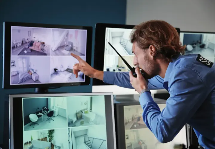 Un guardia de seguridad revisa las imágenes de una cámara de seguridad en varios monitores, comunicándose por radio.