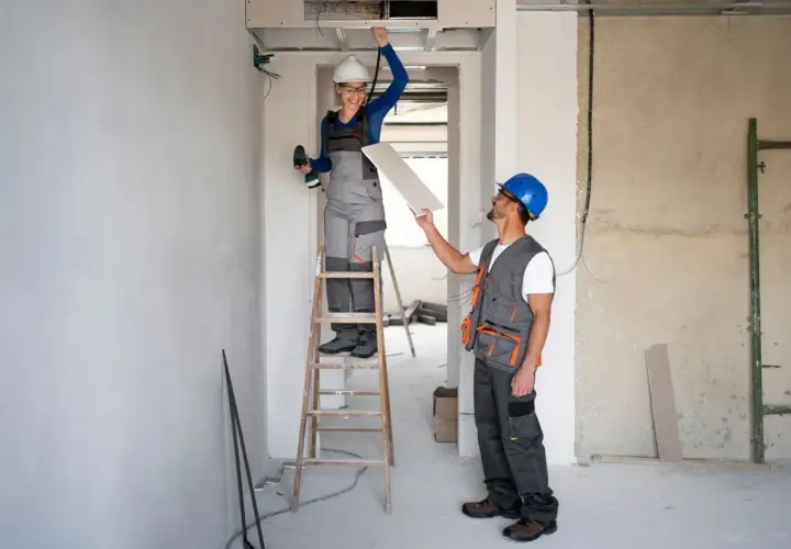 Dwóch budowlańców w kaskach pracuje przy suficie. Kobieta na drabinie trzyma wiertarkę, mężczyzna podaje jej płytę gipsowo-kartonową. W tle widać instalacje i materiały budowlane, jakby przygotowywali się do montażu **fizeliny**.