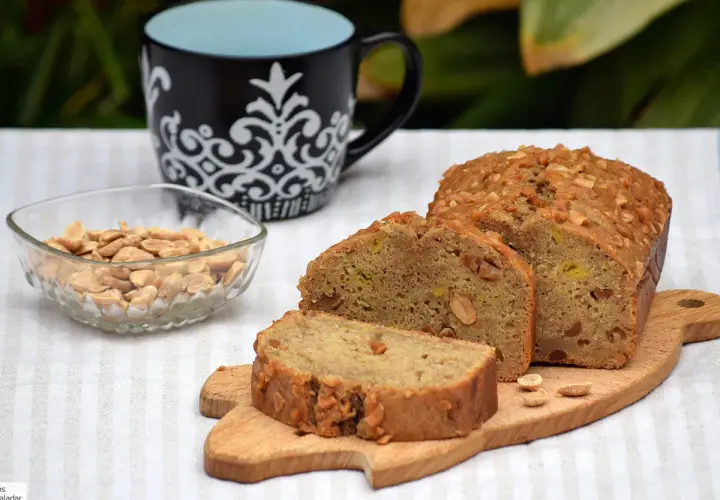 Un delicioso bizcocho de plátano, cubierto de frutos secos, acompañado de cacahuetes y una taza.