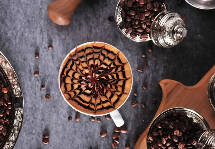 Kawa mocha z artystycznym wzorem na piance. Obok ziarna kawy w ozdobnych naczyniach.
