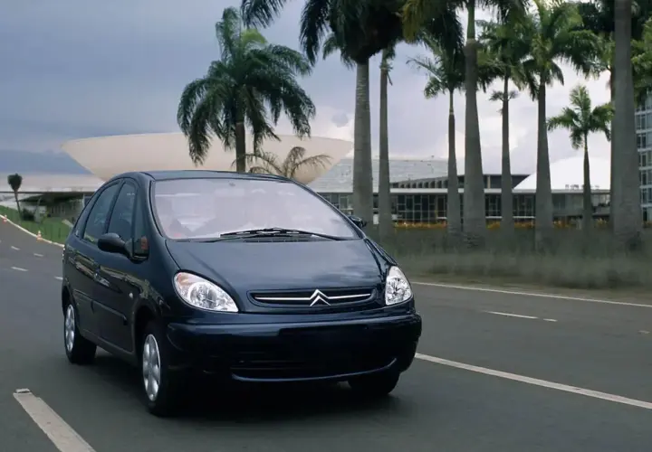 Ciemnoniebieski Citroen Xsara Picasso jedzie drogą wśród palm, w tle nowoczesna architektura.