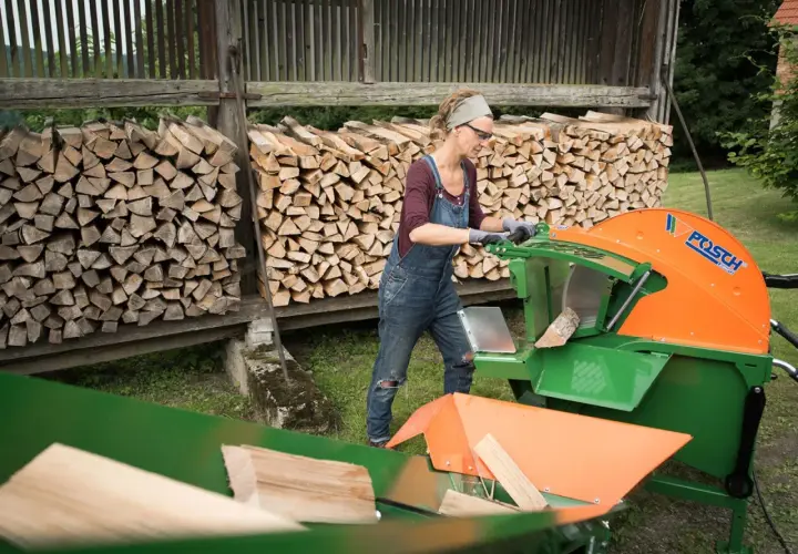Frau bedient eine Schiebetisch Kreissäge für Brennholz, um Holzscheite zu schneiden. Im Hintergrund sind Holzstapel zu sehen.