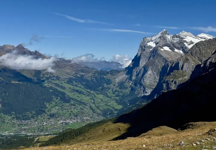 Panoramic view of a valley village nestled among green hills, with dramatic, snow-capped peaks of the Swiss Alps in the background. This scene exemplifies the best hikes in Swiss Alps.