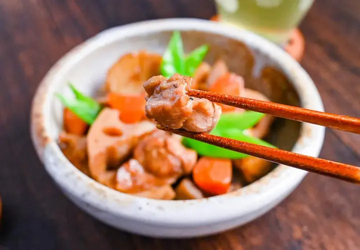 A close-up of a bowl of chikuzenni recipe, with chopsticks lifting a piece of chicken. The dish contains chicken, lotus root, carrots, and green beans.