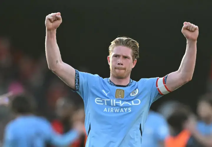 Kevin De Bruyne w jasnoniebieskiej koszulce z podniesionymi rękami, świętujący zwycięstwo.