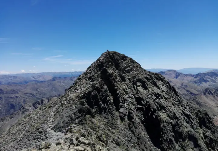 Cima rocosa de montaña con excursionistas y vista panorámica de picos bajo un cielo azul.