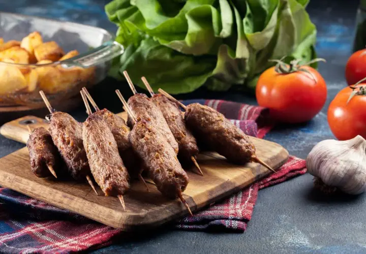 Grillowane ćevapi na drewnianej desce, obok pomidorów, czosnku i pieczonych ziemniaków.