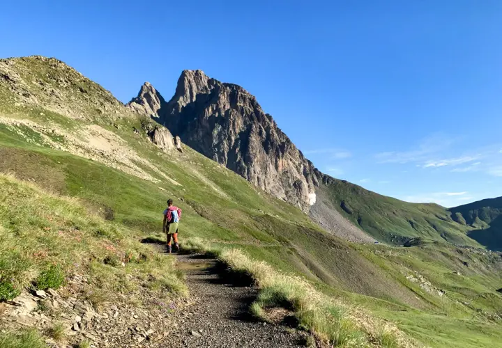 Senderista se dirige hacia el imponente Midi d'Ossau, rodeado de verdes laderas y un cielo azul despejado.