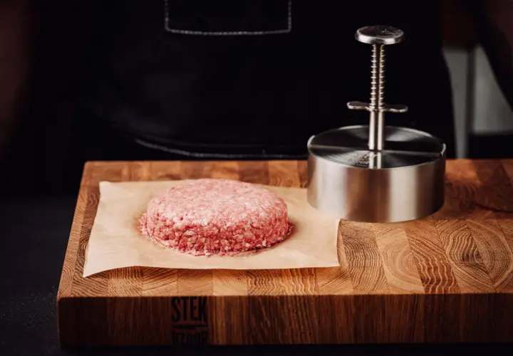 Surowy kotlet na desce i praska. Dowiedz się, dlaczego sól wyciąga wodę z mięsa i kiedy najlepiej solić steki.