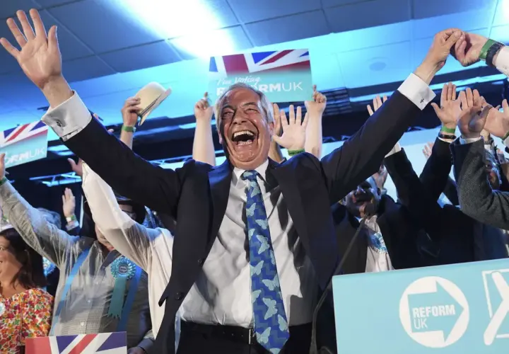 Jubelnde Menge bei einer Wahl in England. Ein Mann mit erhobenen Armen und einem breiten Lachen im Vordergrund, umgeben von Union-Jack-Flaggen und "Reform UK"-Schildern.