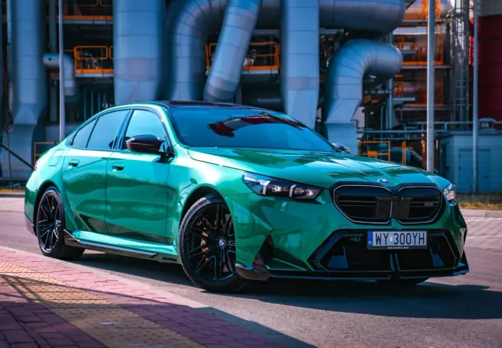 Błyszczące, zielone BMW M5 G90 na tle przemysłowej infrastruktury.