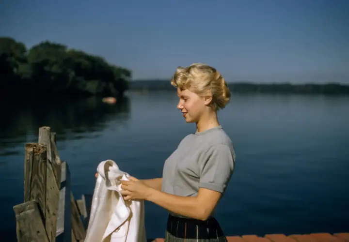 Joven con peinados años 50, con un suéter gris y falda a cuadros, sostiene una chaqueta blanca junto a un lago.