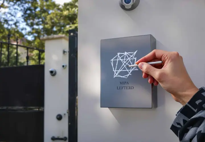 Main MIPA LEFTERD, une main trace un motif géométrique sur une plaque. Une caméra de surveillance est visible au-dessus, comme un marquage cambriolage discret.