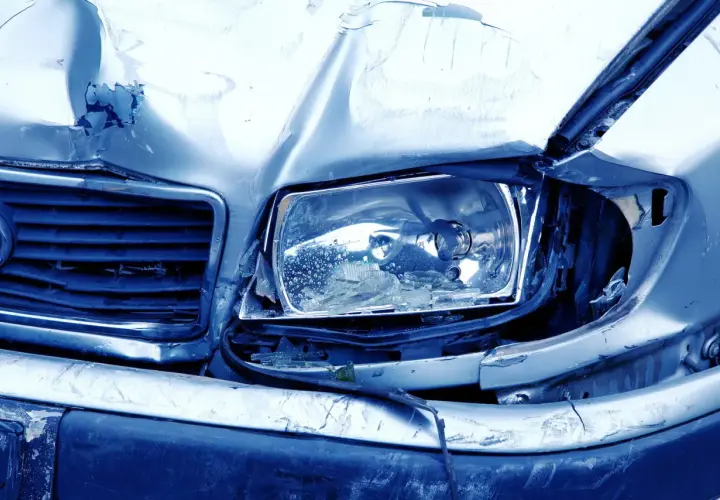Il cofano e il faro di un'auto sono gravemente danneggiati, con lamiera accartocciata e vetro rotto. Sembra il risultato di **atti vandalici**.