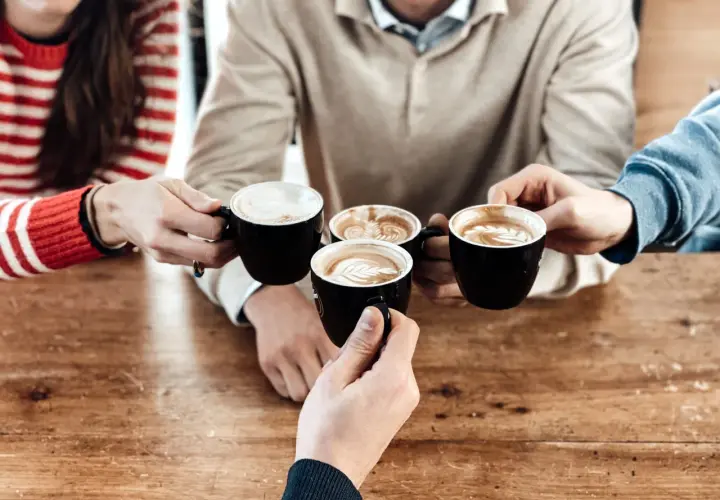 Un brindisi con caffè al bar con amici. Quattro tazze nere con schiuma artistica si avvicinano su un tavolo di legno.