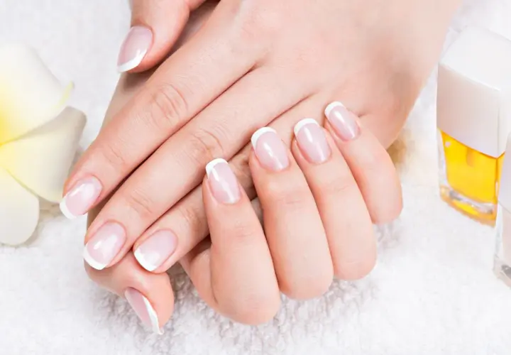 Elegancki manicure french z diamencikami, dłonie zadbane i wypielęgnowane, obok kwiat i lakiery.
