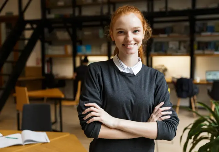 Uśmiechnięta studentka filologii hiszpańskiej z rudymi włosami, w bibliotece z książkami w tle.