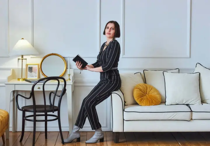 Kobieta w eleganckim, prążkowanym stroju siedzi na kanapie, trzymając książkę. To kwintesencja stylu glamour – wyrafinowanie i blask.