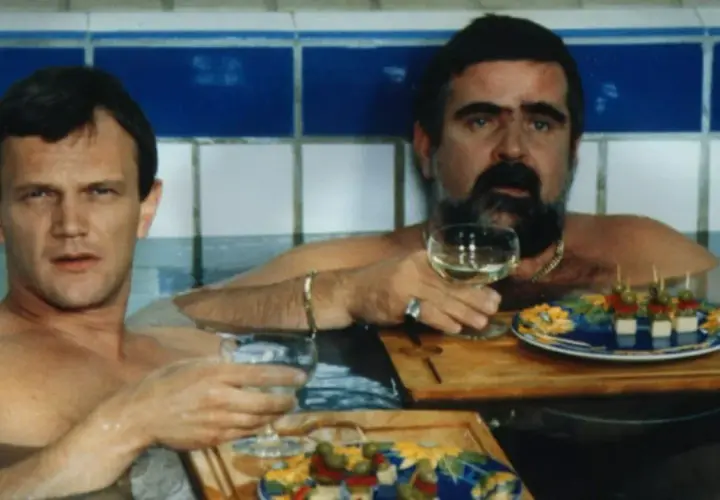 Dwóch mężczyzn w wannie, jeden z wąsami, pije wino i je przekąski. Scena z filmu "Kiler", rok produkcji 1997.