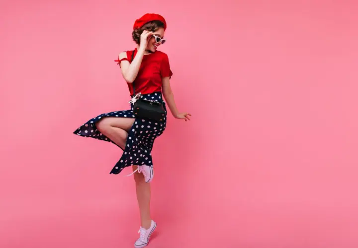 Dziewczyna w stylu pin-up w czerwonej bluzce i spódnicy w grochy tańczy na różowym tle.
