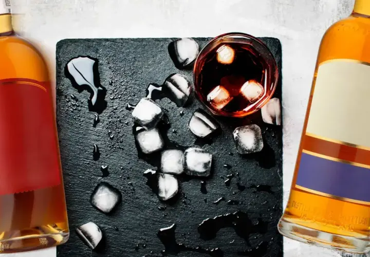 Dwie butelki whisky, szklanka z lodem i rozlaną wodą. Metaliczny posmak i pieczenie to objawy niskiej jakości whisky zbożowej.