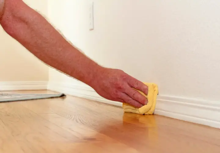 Mano limpiando rodapiés con un paño amarillo. El suelo de madera brilla y la pared está pintada de blanco.