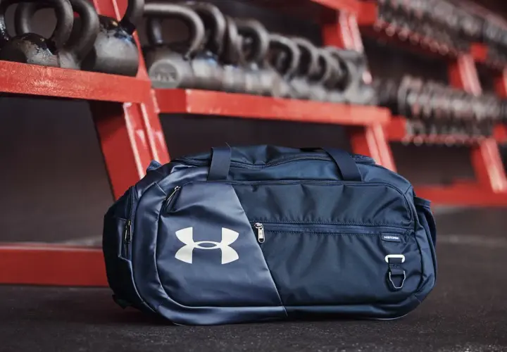 Granatowa torba sportowa 100l z logo Under Armour, gotowa na trening. W tle sztangi i hantle.