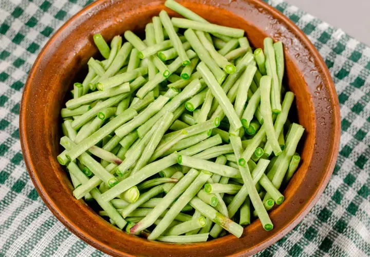 Fagiolini in umido freschi e verdi, pronti per essere cucinati in una ciotola di terracotta su una tovaglia a quadretti.