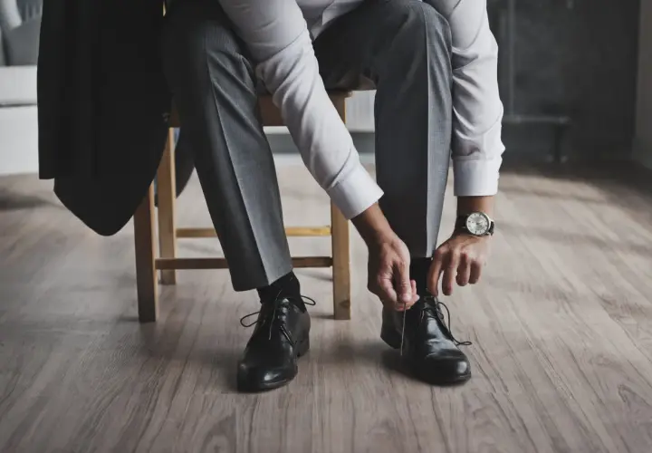 Mężczyzna w garniturze, z marynarką na krześle, zawiązuje buty. Elegancki, śniady kolor stroju i klasyczne obuwie.