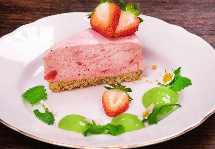 En bit krämig cheesecake jordgubb med färska bär och mynta på en vit tallrik.