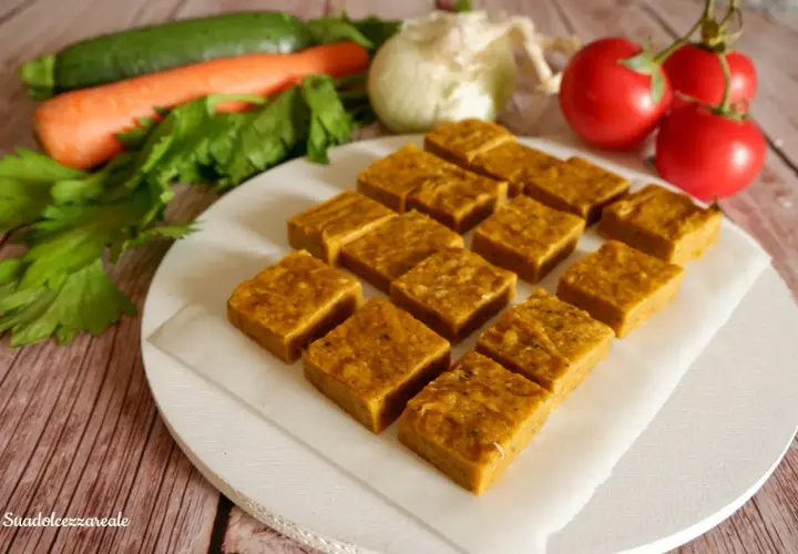 Cubetti di dado vegetale fatto in casa, pronti per insaporire ogni piatto. Verdure fresche come carote, zucchine e pomodori sullo sfondo.