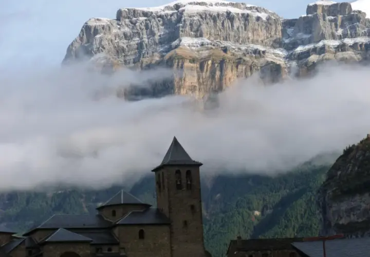 Iglesia románica con campanario, rodeada de niebla, bajo imponentes montañas nevadas. Ideal para explorar rutas desde Torla.