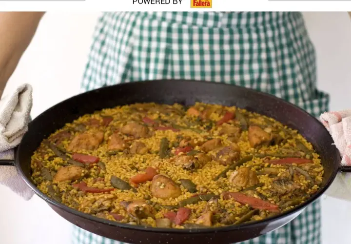 Una persona con un delantal a cuadros verdes sostiene una paella valenciana recién hecha, llena de arroz, pollo, judías verdes y pimientos rojos.