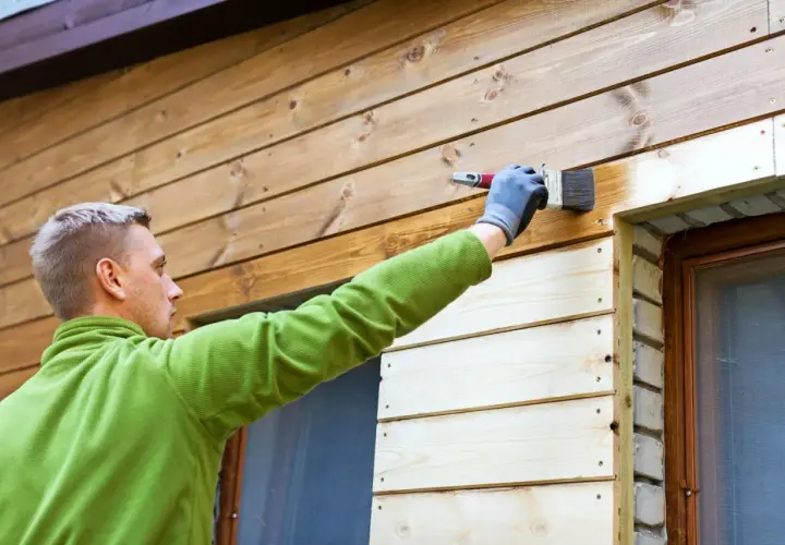 Man målar husfasaden med en pensel. Träet är ljusbrunt och ser ut att vara tryckimpregnerat.