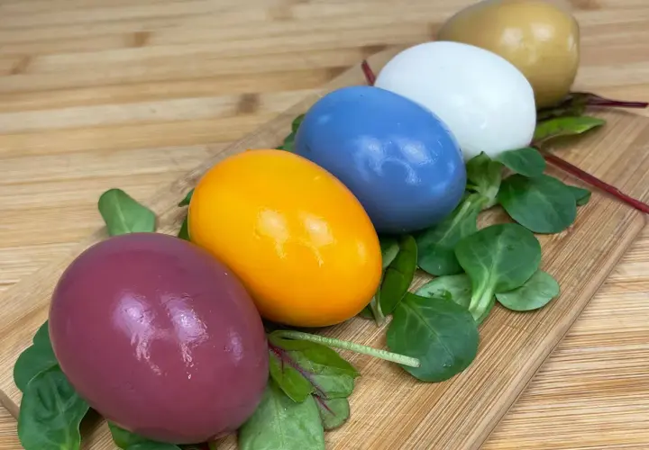 Cinq œufs colorés, du violet à l'orange vif, en passant par le bleu, le blanc et le doré, disposés sur un lit de verdure sur une planche en bois. Chaque couleur oeuf est une surprise.