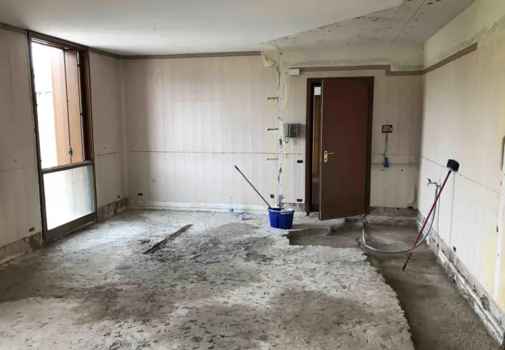 Interno di una stanza in ristrutturazione, con il pavimento in cemento grezzo e pareti spoglie. Si notano attrezzi da lavoro, tra cui un secchio blu e una scopa, che suggeriscono i lavori in corso e i possibili svantaggi dell'intonaco armato.