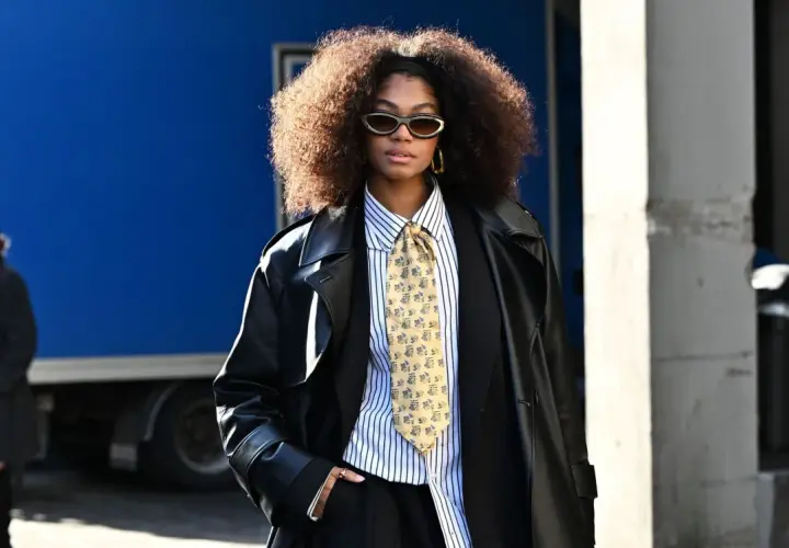 Une femme porte un pantalon noir, une chemise à rayures, une cravate fantaisie et un long manteau en cuir. Ses bottines sont cachées sous son pantalon.