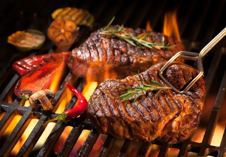Deux steaks barbecue juteux, ornés de romarin, grésillent sur un barbecue avec des poivrons rouges, des champignons et des piments.