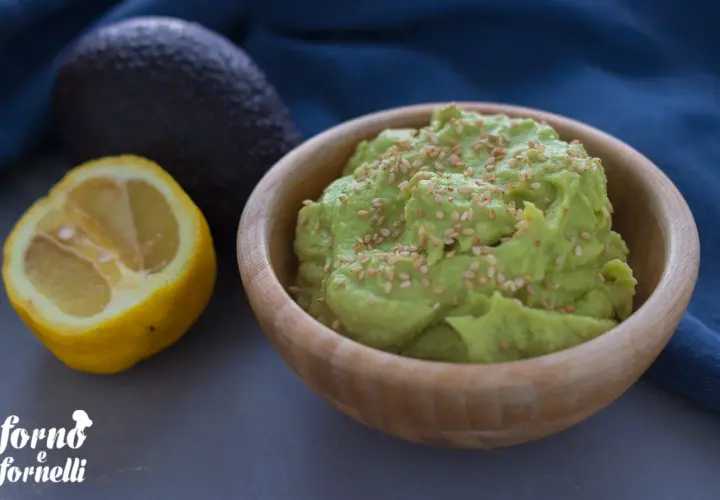 Hummus di avocado cremoso in ciotola di legno, guarnito con semi di sesamo. Accanto, un avocado intero e mezzo limone.