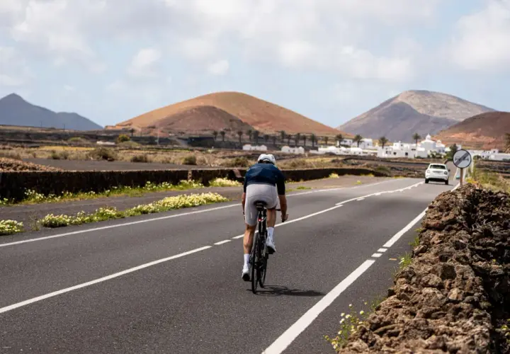 Ciclista pedaleando por una carretera en Lanzarote, con volcanes y casas blancas al fondo. Una aventura de lanzarote en bici.