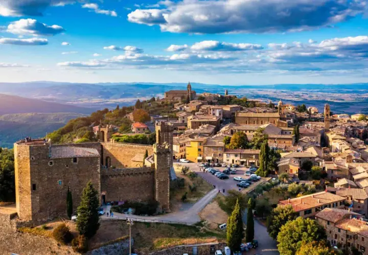 A panoramic view of medieval towns in Italy, perched on a hilltop under a bright blue sky with fluffy clouds.