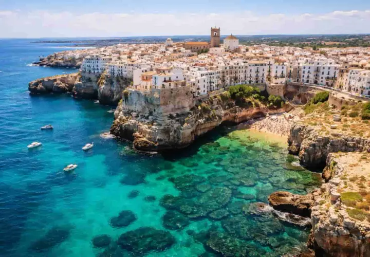 Vue aérienne de Polignano a Mare, avec ses maisons blanches perchées sur des falaises rocheuses surplombant une mer turquoise et une plage bondée.