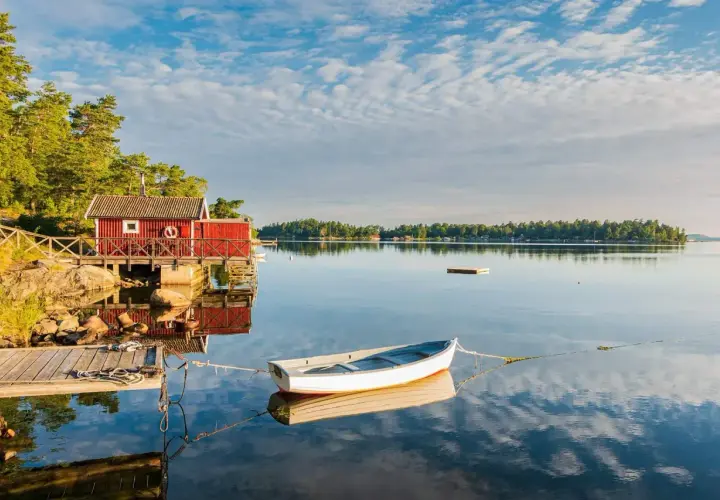 En vit båt ligger förtöjd vid en röd sjöbod. Kanske är det en båt till Drottningholm, redo för en lugn tur på vattnet.