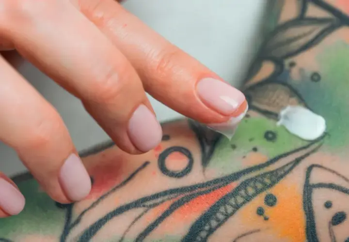 Main d'une personne appliquant une crème sur un tatouage coloré. Si vous suspectez une infection tatouage, que faire ? Consulter un professionnel de santé.