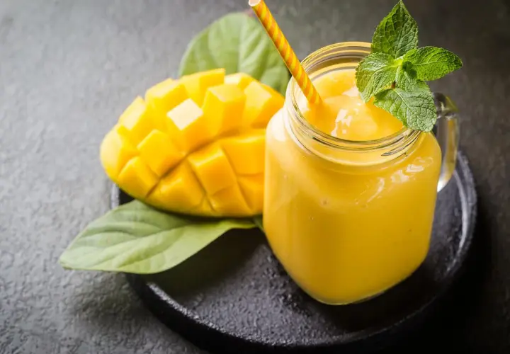 Un smoothie mangue onctueux dans un pot en verre, garni de menthe fraîche, à côté d'une moitié de mangue coupée en dés.