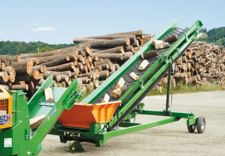 Grünes Förderband transportiert Holzscheite. Im Hintergrund liegt ein großer Stapel Baumstämme.