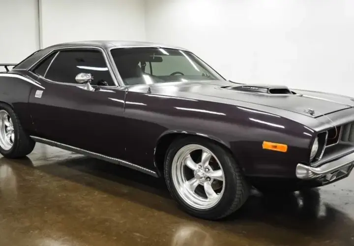 Klasyczny Plymouth Barracuda w ciemnym kolorze, z chromowanymi felgami i sportowym wlotem powietrza na masce.