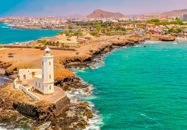 Faro blanco en una costa rocosa con aguas turquesas. Un pueblo costero se extiende detrás. ¡Ideas de qué hacer en Cabo Verde!